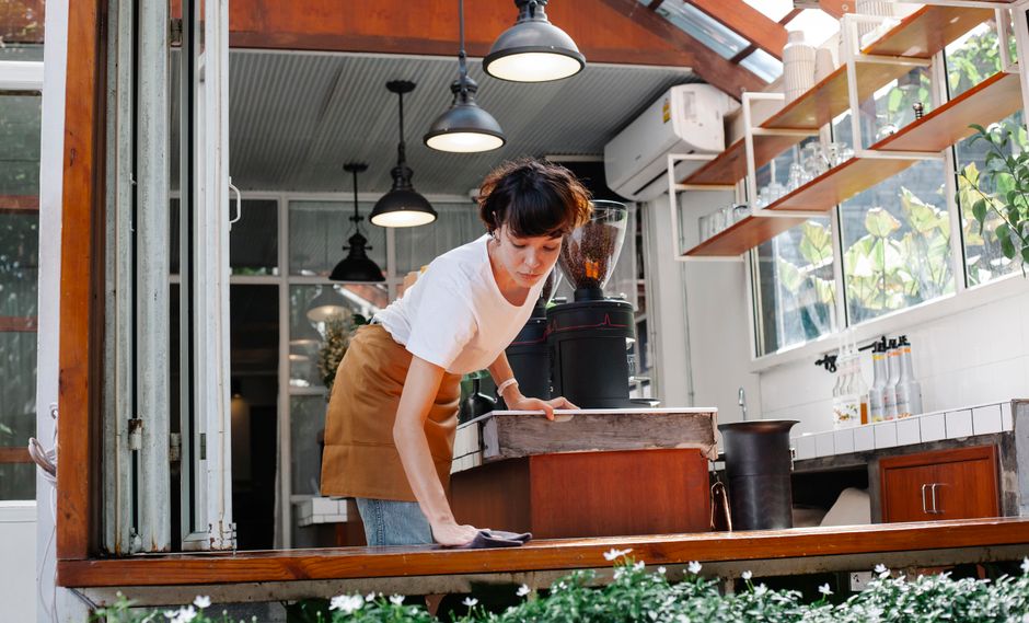 A woman performing surface cleaning in a bright, modern café or small shop, wearing a white t-shirt and a tan apron. She is using a cloth to wipe a wooden counter, which appears clean and polished. T