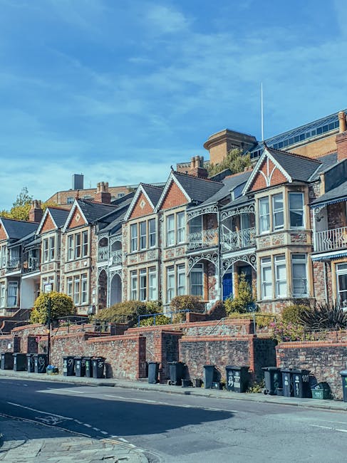 A residential street in Barnes Village featuring a row of red-brick houses with pitched roofs, some with white window frames and small front gardens. In the foreground, a silver compact car is parked 